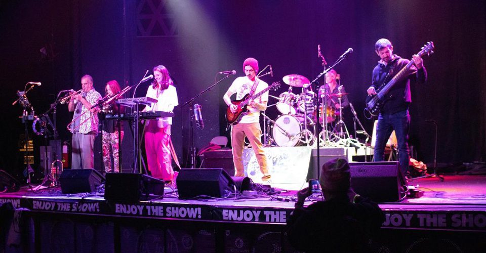 A six-member band performs on stage with various instruments including a saxophone, keyboard, guitar, drums, and bass. Stage lights and audience members are visible.