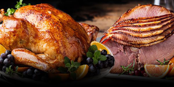 Roasted turkey and sliced ham are displayed with blueberries, herbs, and orange slices on a rustic table.