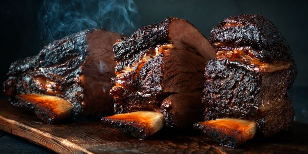 Three large, smoked beef short ribs with a dark, caramelized crust are arranged on a wooden board, with visible bone and steam rising from the meat.
