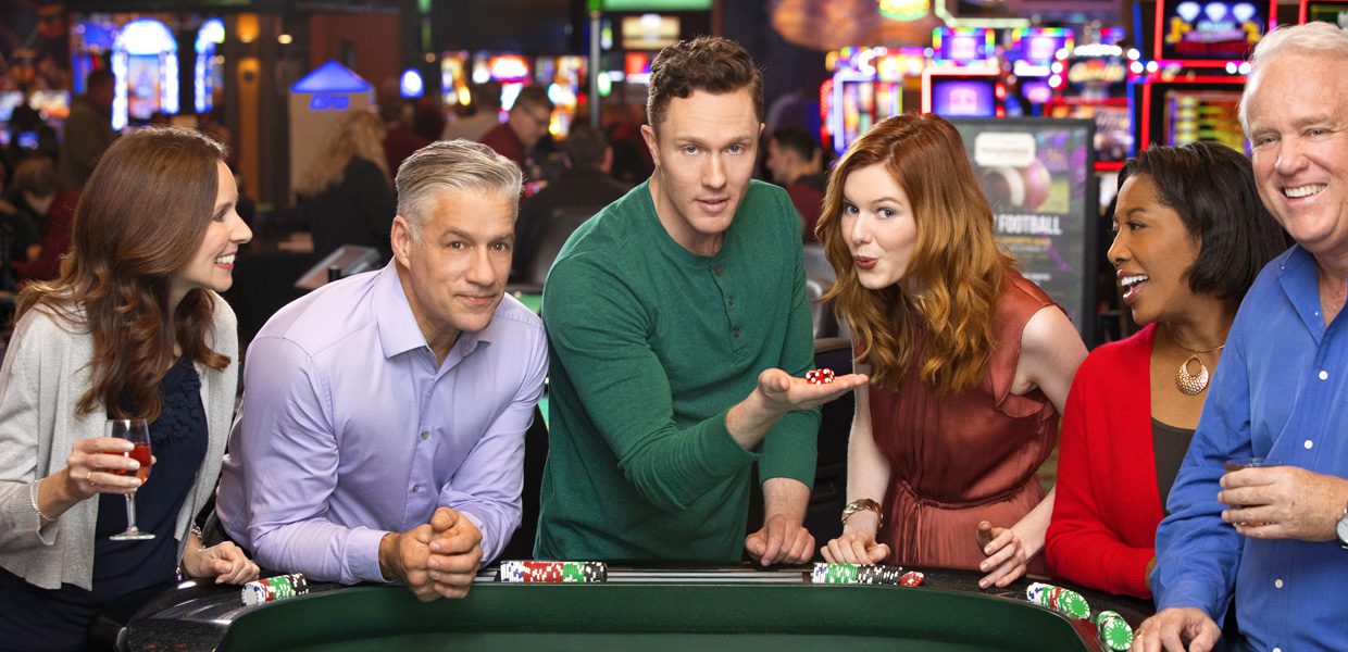 Group of six people gathered around a casino table, smiling and holding poker chips. Background features colorful casino lights and slot machines.