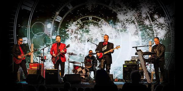 A five-member band performs on stage with guitars, drums, and keyboard. The backdrop features a clock design and bright, firework-like lights.