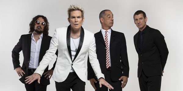 Four men in suits stand against a plain white background; the man in front wears a white blazer, while the others wear black.
