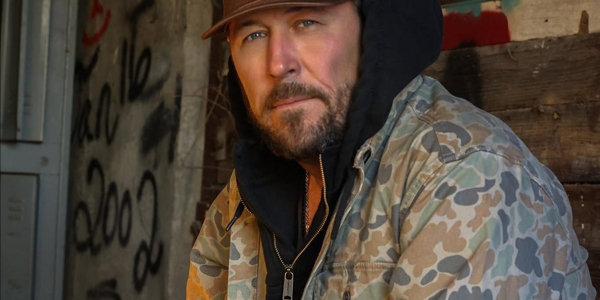 A man in a camouflage jacket and brown cap sits indoors near a wooden wall with graffiti in the background.