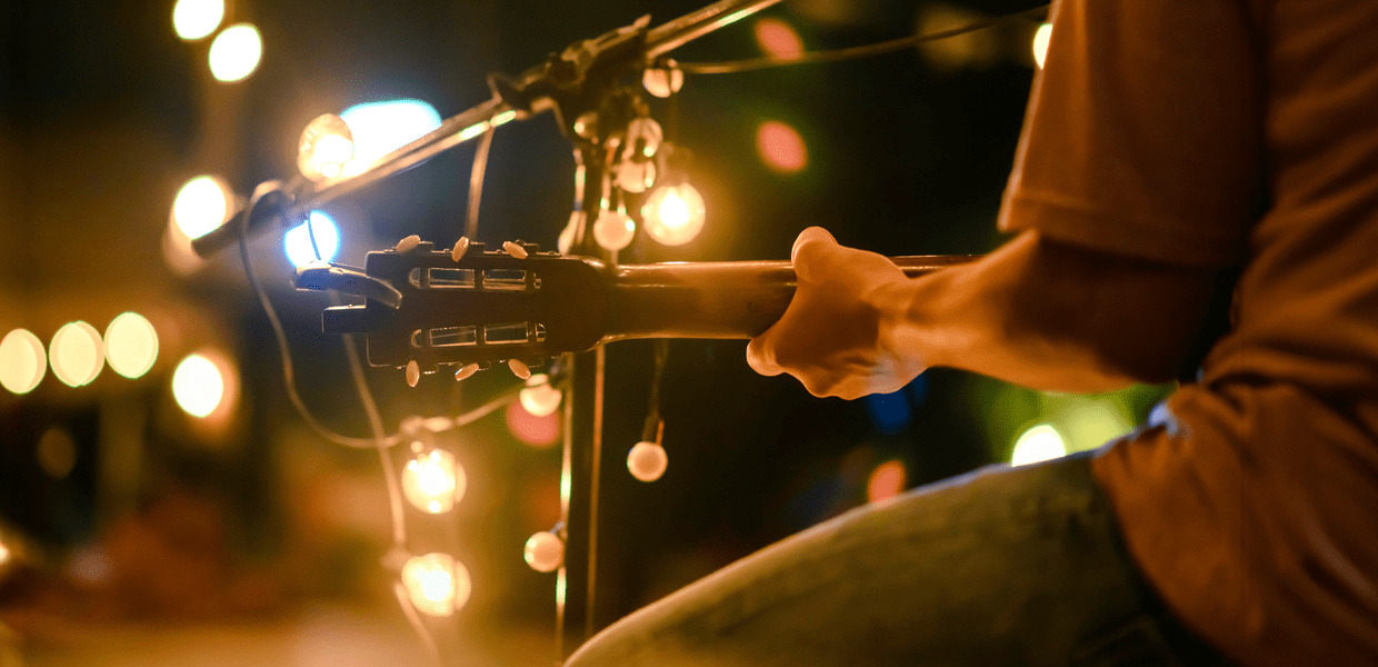 A person plays an acoustic guitar on stage, decorated with string lights, with the focus on the guitar and the person's arm.